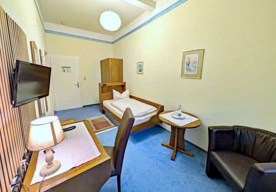 Kleinzimmer mit Einzelbett, Fernseher, Tisch, Stuhl und gemütlicher Sitzecke.