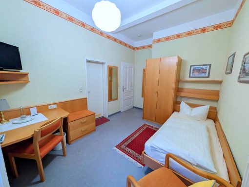 Ein einfaches Zimmer mit Einzelbett, Schreibtisch und Fernseher. Helle Wände und Möbel.