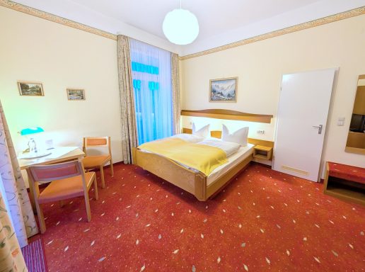 Helles Hotelzimmer mit Doppelbett, Tisch, Stühlen und Balkon. Rote Teppichboden.