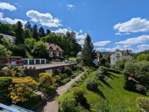 Landschaft mit grünen Gärten und idyllischen Häuschen unter blauem Himmel.