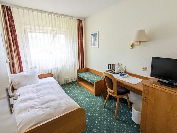 Helles Einzelzimmer mit Schreibtisch, TV und Fenster im Hintergrund.
