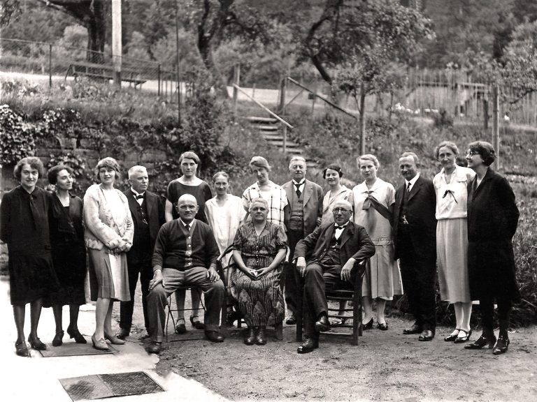 Eine Gruppe von Menschen steht und sitzt in einem Garten, teils in formeller Kleidung. 1930er Jahre