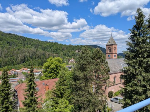Panoramablick auf eine Stadt mit Kirche, umgeben von grünen Hügeln und Wolken.