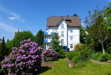 Weißes Haus mit blauen Balkonen, umgeben von lila Blumen und grünem Garten.