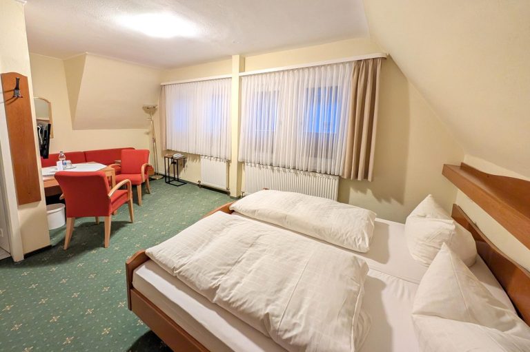Geräumiges Hotelzimmer mit zwei Betten, Tisch und roten Stühlen. Helle Vorhänge.