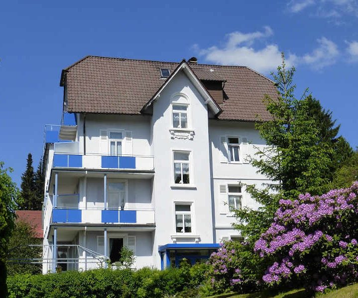 Helles mehrstöckiges Haus mit Balkon, umgeben von Bäumen und blühenden Pflanzen.