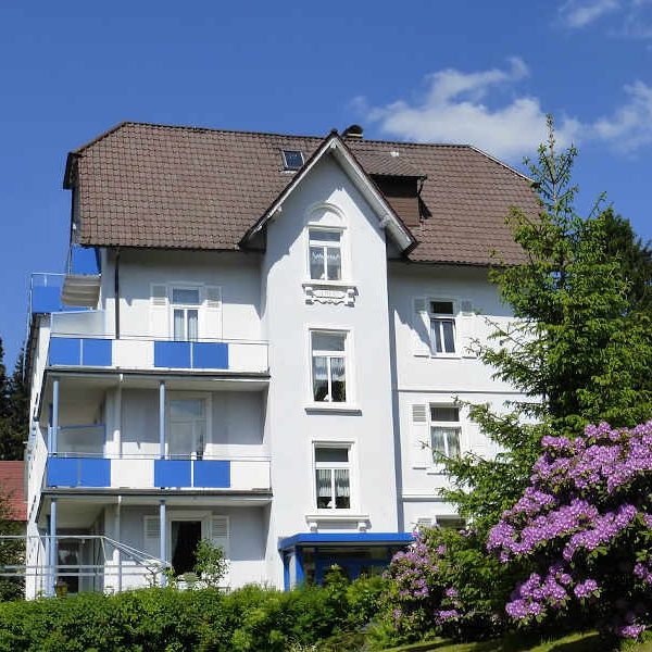 Weißes Haus mit blauen Balkonen, umgeben von Bäumen und blühenden Sträuchern.