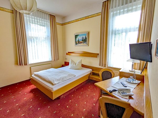 Helles Hotelzimmer mit Doppelbett, Tisch, Stuhl und Fernseher. Rote Teppichboden.