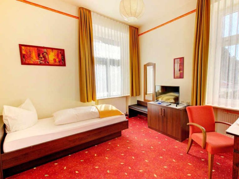 Ein einfaches Hotelzimmer mit Einzelbett, Schreibtisch, Stuhl und Fenster mit Vorhängen.