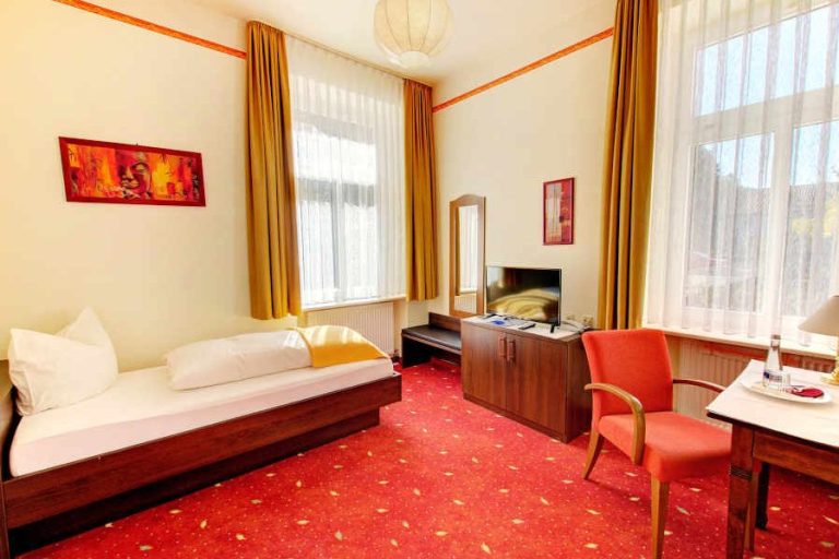 Helles Hotelzimmer mit Einzelbett, Schreibtisch und Fernseher. Rote Teppich und Vorhänge.