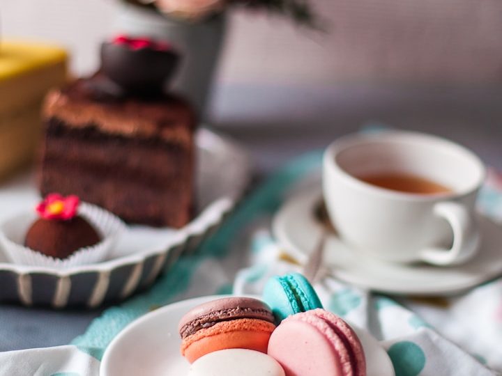 Bunte Macarons, eine Schokoladentorte und eine Tasse Tee auf einem gedeckten Tisch.