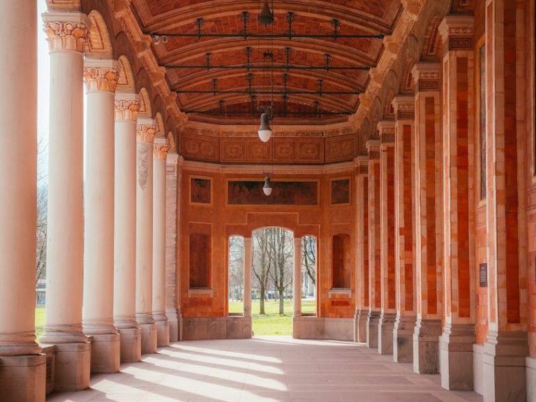 Säulenhalle mit roten Wänden und Sicht auf einen Park im Hintergrund.