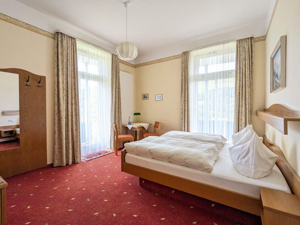 Helle Hotelzimmer mit Doppelbett, Tisch, Stühlen und Balkon. Rote Teppichboden.