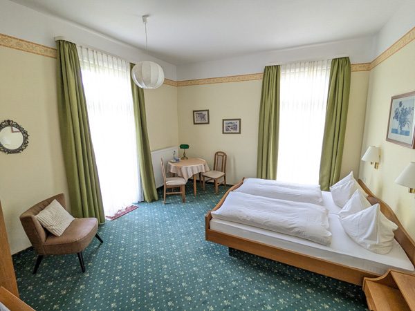 Helles Zimmer mit Doppelbett, Tisch, Stühlen und großen Fenstern mit Grünen Vorhängen.