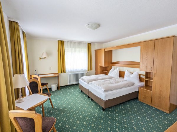 Helles Hotelzimmer mit Doppelbett, Tisch, Stuhl und Holzregal sowie grünen Teppichboden.