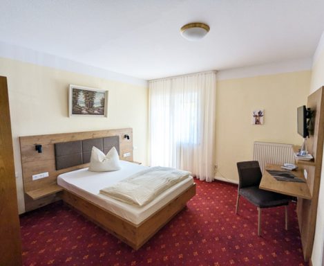 Helles Gästezimmer mit Doppelbett, Schreibtisch und Stuhl, rotem Teppichboden.