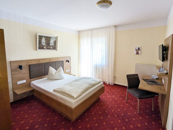 Gemütliches Einzelzimmer mit Tisch, Stuhl und Fenstern, dekoriert in warmen Farben.