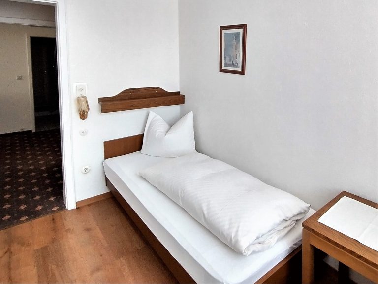 Ein einfaches Einzelzimmer mit einem Bett, Nachttisch und einem Bild an der Wand.