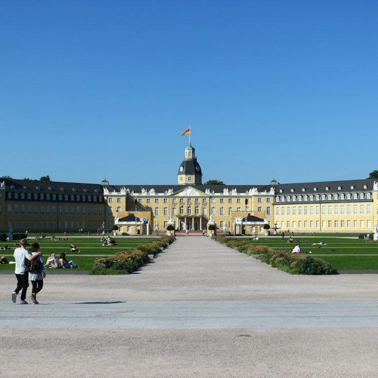 Schloss Karlsruhe mit Blumenbeeten und Spaziergängern im Vordergrund, blauer Himmel.